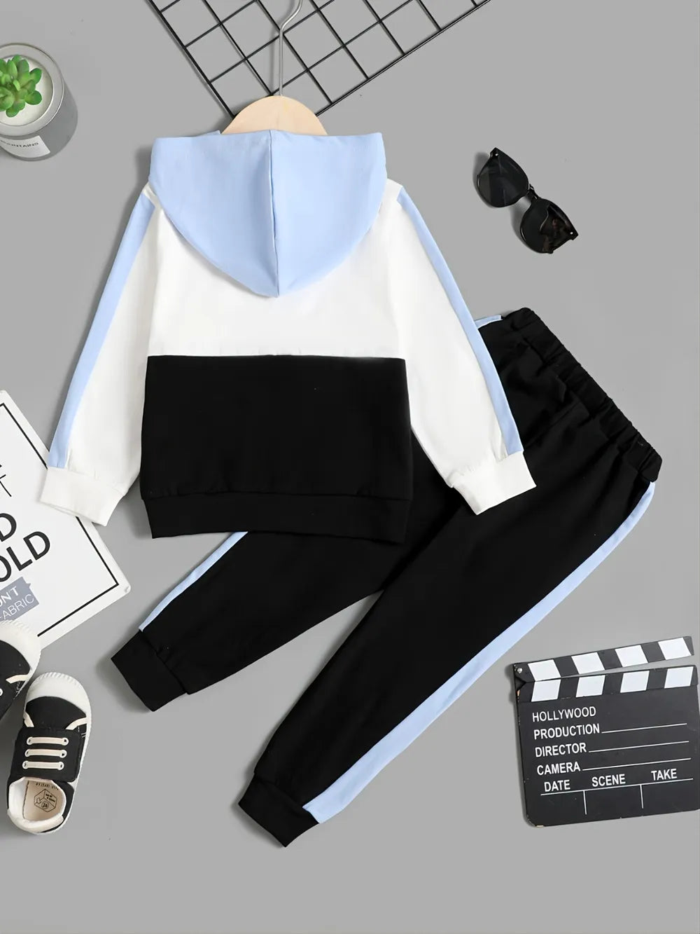 Two-tone hoodie and black pants set on a gray surface with a clapperboard and sneaker in the background.