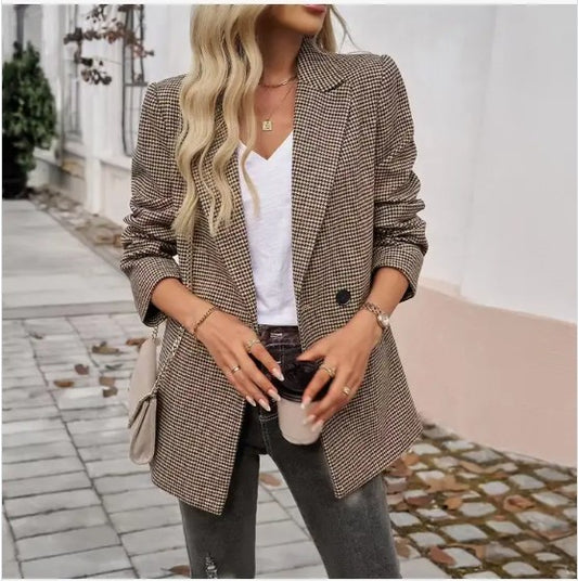 A lady in a brown Houndstooth blazer with a white top, grey jeans, and some dope accessories like a beige purse and gold jewelry. She's posing on a stylish cobblestone sidewalk with a white wall backdrop.