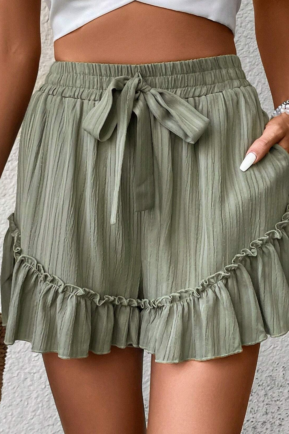 A woman wearing high-waisted, light green shorts with a drawstring waist and ruffled hem. She is dressed casually with a crop top, and her hand is resting on her hip, standing against a textured background.