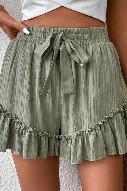 A woman wearing high-waisted, light green shorts with a drawstring waist and ruffled hem. She is dressed casually with a crop top, and her hand is resting on her hip, standing against a textured background.