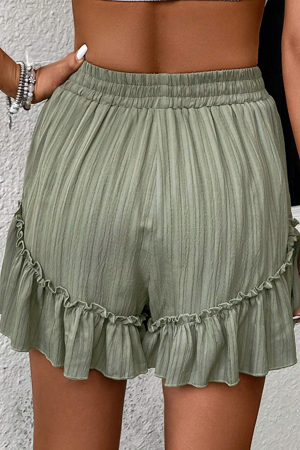 A woman wearing high-waisted, light green shorts with a drawstring waist and ruffled hem. She is dressed casually with a crop top, and her hand is resting on her hip, standing against a textured background.