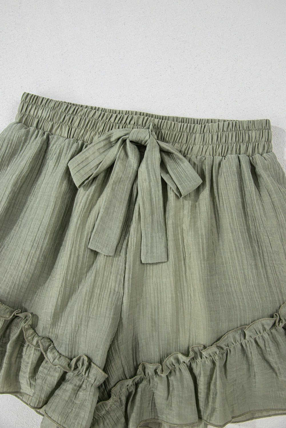 A close-up of light green, textured shorts with a drawstring waist and ruffled hem, displayed against a neutral background.
