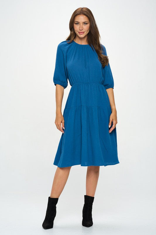 Woman wearing a cotton tiered midi dress with ¾ sleeves and a gathered waist, styled with black crisscross heels against a white background.