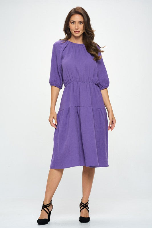 Woman wearing a purple tiered midi dress with ¾ sleeves and a gathered waist, styled with black crisscross heels against a white background.