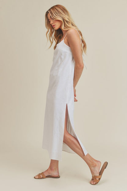 Woman in a vibrant white dress striking a pose against a neutral beige background. The dress features thin straps and a side slit, complemented by her long blonde hair and silver jewelry.
