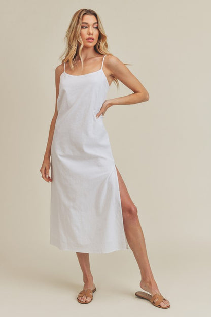 Woman in a vibrant white dress striking a pose against a neutral beige background. The dress features thin straps and a side slit, complemented by her long blonde hair and silver jewelry.