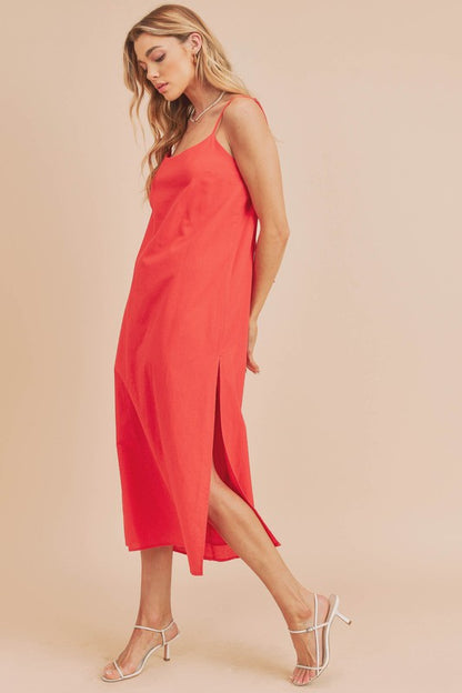 Woman in a vibrant red dress striking a pose against a neutral beige background. The dress features thin straps and a side slit, complemented by her long blonde hair and silver jewelry.