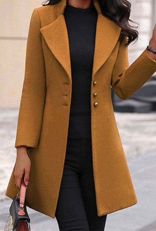 The image shows a woman wearing a long, camel-colored coat with big lapels and brass buttons over a black top and pants. She's holding a purse in one hand and has long, dark hair.