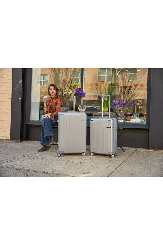 Two silver hard-shell suitcases with four wheels sit in front of a storefront window, beside a person on a bench. Reflections of trees and buildings suggest an urban setting.