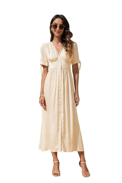 A woman in a cream-colored maxi dress with short sleeves and a V-neckline, wearing sunglasses and strappy heels, standing against a white background.

