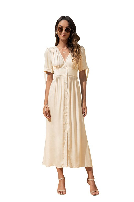A woman in a cream-colored maxi dress with short sleeves and a V-neckline, wearing sunglasses and strappy heels, standing against a white background.

