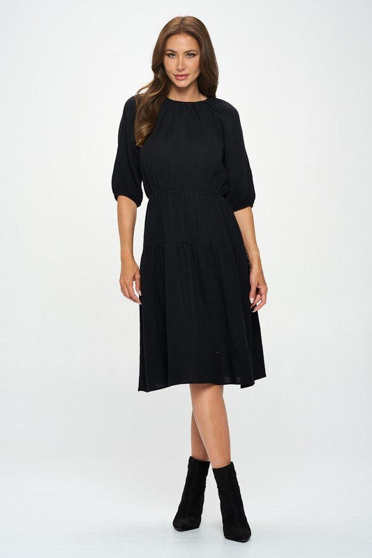 Woman wearing a black midi dress with elbow-length sleeves, gathered waist, and modest flare skirt, styled with black ankle boots.