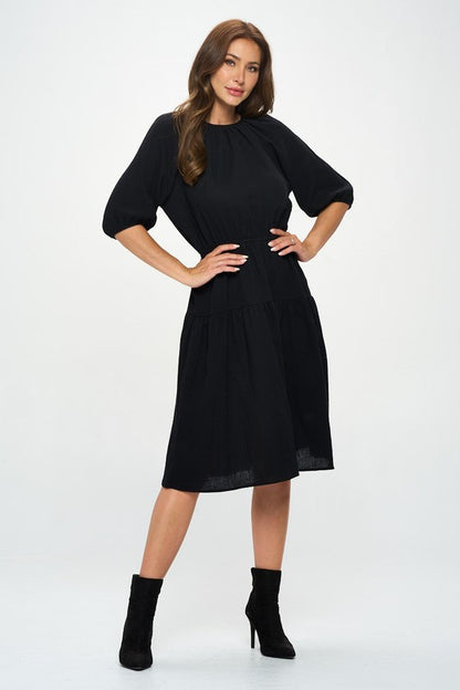 Woman wearing a black midi dress with elbow-length sleeves, gathered waist, and modest flare skirt, styled with black ankle boots.