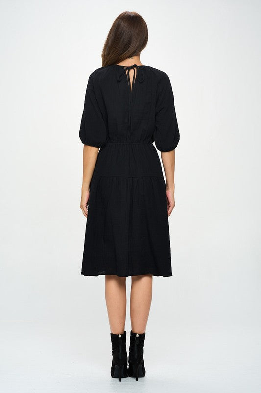 Woman wearing a black midi dress with elbow-length sleeves, gathered waist, and modest flare skirt, styled with black ankle boots.