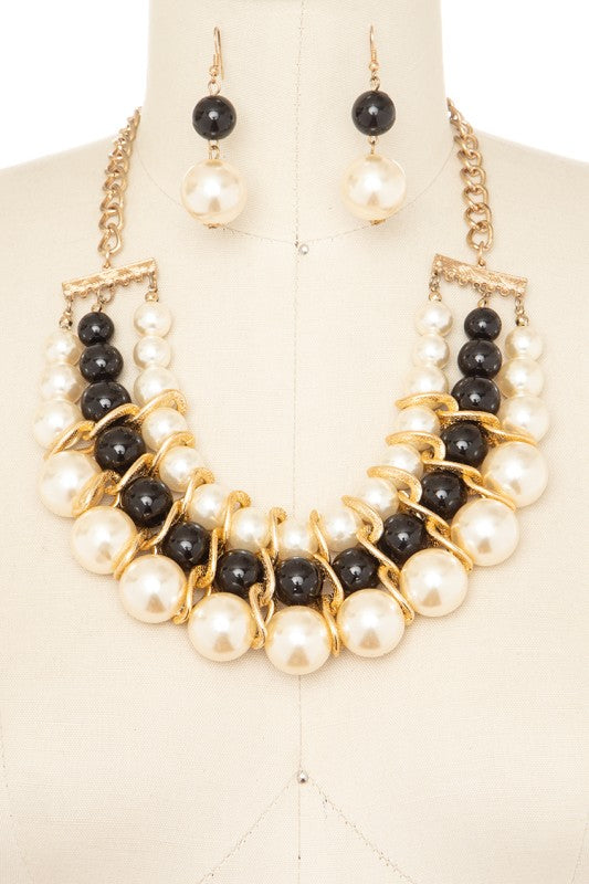 A necklace and earring set on a mannequin. The necklace has gold chains attaching to a bar with black and white pearls wrapped in gold wire. The earrings are black and white pearls hanging from gold hooks.