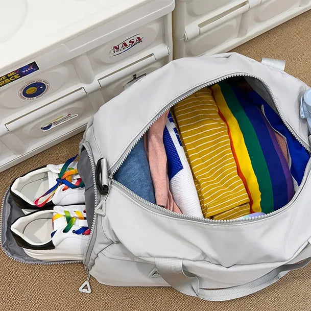 Open light gray duffel bag on a carpeted floor, neatly packed with colorful clothes including striped shirts and a pair of white sneakers with rainbow laces in a separate compartment. NASA-themed stickers on nearby drawers add a playful, space-inspired touch—perfect for organized travel prep.