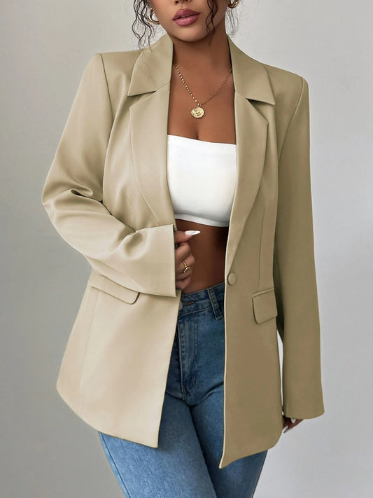 A woman wearing a tan blazer, a white crop top, and blue jeans. She's posing with one hand on her hip, showing off some stylish accessories like a gold necklace and rings. The background is a plain gray wall.