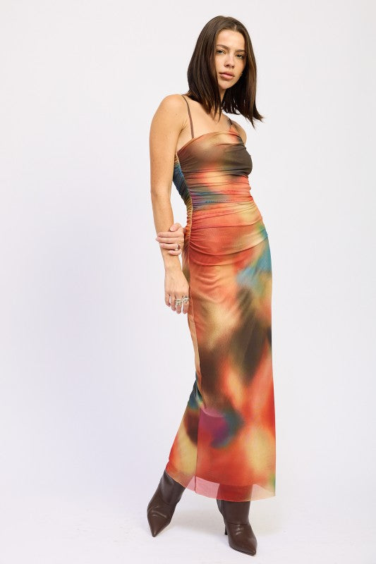 A woman wearing a vibrant, multicolored one-shoulder maxi dress with brown boots, posing against a plain white background.