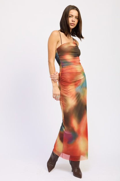 A woman wearing a vibrant, multicolored one-shoulder maxi dress with brown boots, posing against a plain white background.