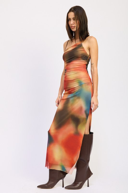 A woman wearing a vibrant, multicolored one-shoulder maxi dress with brown boots, posing against a plain white background.
