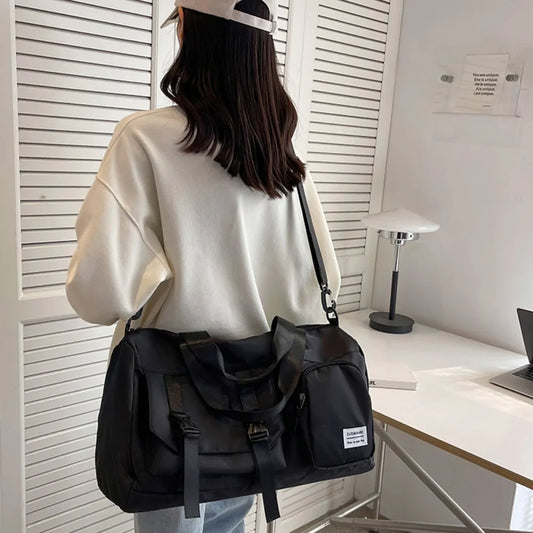 A woman in a white oversized sweatshirt and light-colored cap stands indoors, carrying a large black duffel bag with a “FASHION MINI GIRL” label.