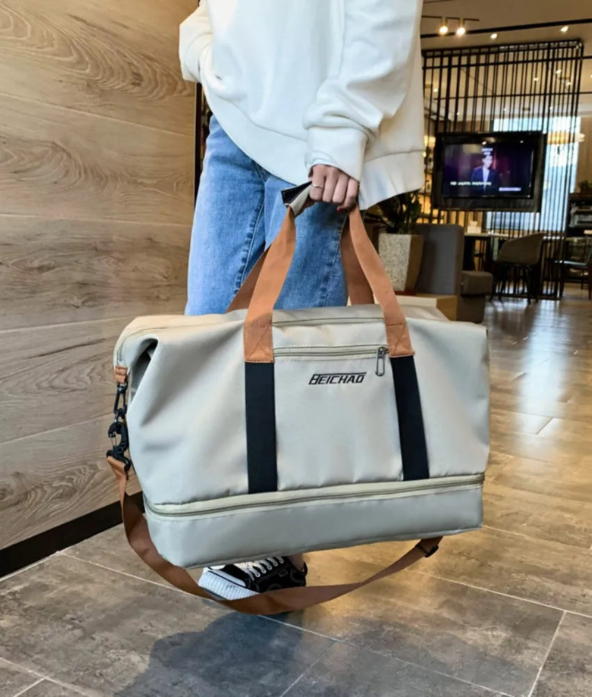 Light gray duffel bag with tan and black straps, featuring multiple compartments and a front. Carried by a casually dressed person outdoors—stylish, spacious, and ideal for travel or gym use.