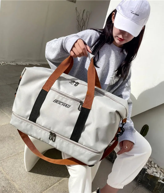 Light gray duffel bag with tan and black straps, featuring multiple compartments and a front. Carried by a casually dressed person outdoors—stylish, spacious, and ideal for travel or gym use.