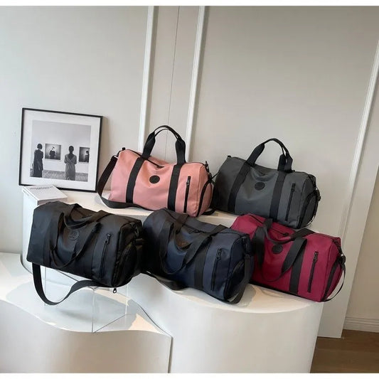 Five cylindrical duffel bags in pink, gray, black, navy blue, and maroon are neatly displayed on a curved white surface in a minimalistic room. Each bag features a circular logo and zippered pocket—stylish and versatile, ideal for travel or gym use.