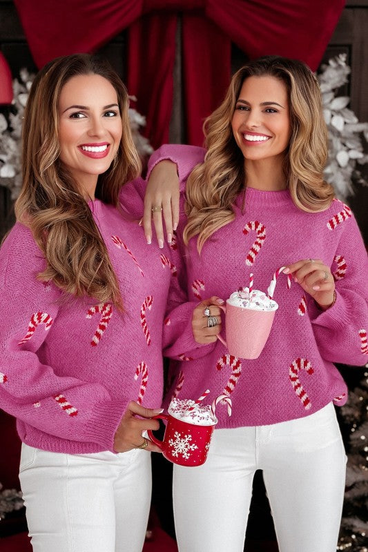 Pair of women wearing pink holiday sweaters with candy cane design and white pants holding seasonal drinks