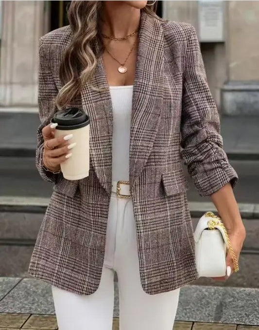 A woman dressed in a stylish outfit, standing outdoors. She's wearing a brown plaid blazer over a white top and pants, accessorized with a white purse, gold belt, and layered necklaces.