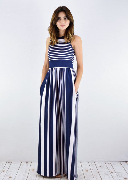 A woman in a blue and white striped maxi dress with a halter neck, posing against a white wall on a wooden floor.