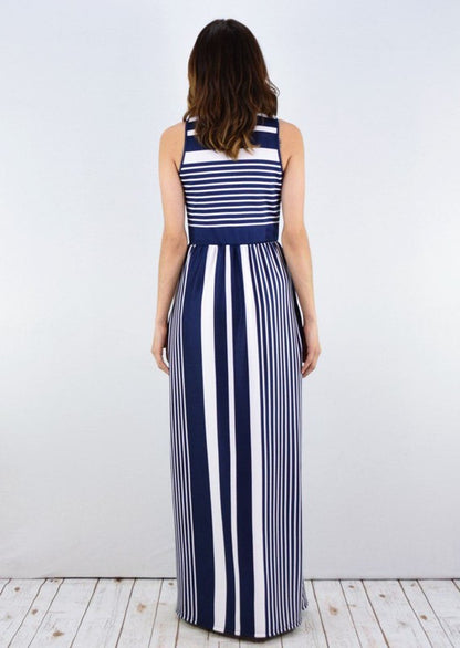 A woman in a blue and white striped maxi dress with a halter neck, posing against a white wall on a wooden floor.