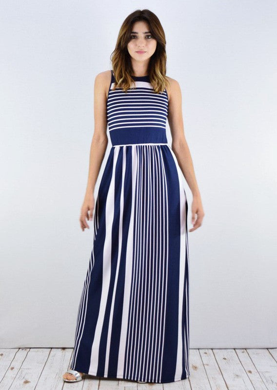 A woman in a blue and white striped maxi dress with a halter neck, posing against a white wall on a wooden floor.