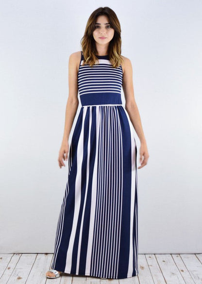 A woman in a blue and white striped maxi dress with a halter neck, posing against a white wall on a wooden floor.