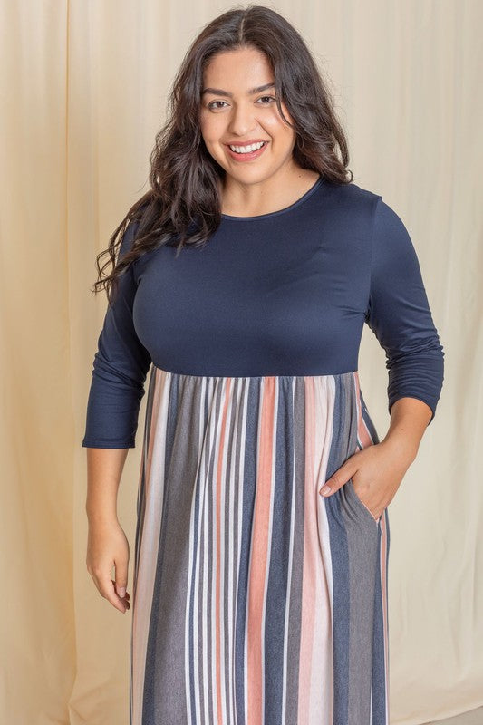 A woman with long dark hair posing in a navy blue top with a striped maxi skirt. The skirt has vertical stripes in shades of blue, pink, white, and gray. 