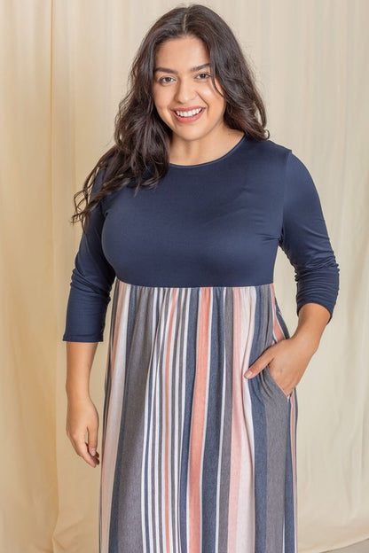 A woman with long dark hair posing in a navy blue top with a striped maxi skirt. The skirt has vertical stripes in shades of blue, pink, white, and gray. 