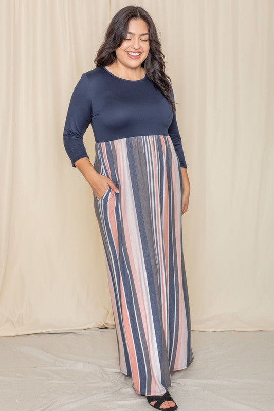 A woman with long dark hair posing in a navy blue top with a striped maxi skirt. The skirt has vertical stripes in shades of blue, pink, white, and gray. 
