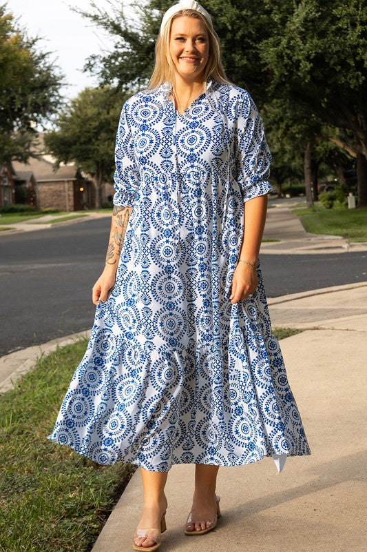  A woman standing on a sidewalk, wearing a blue and white patterned dress with a collar and elbow-length sleeves.