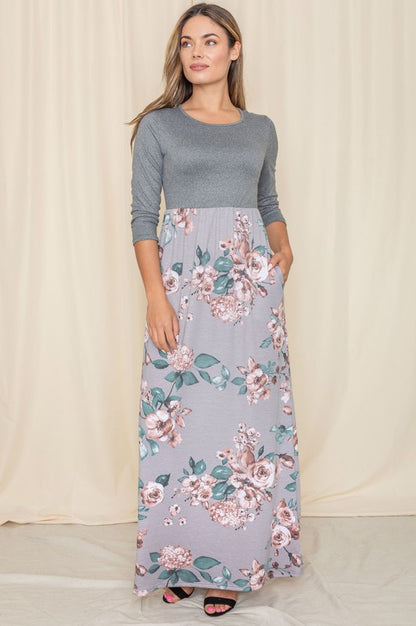 A woman in a long dress with a gray top and a flowy floral skirt stands against a cream-colored curtain backdrop