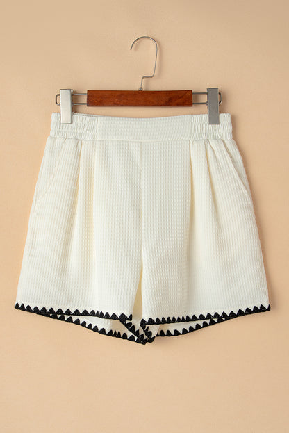 A lady wearing high-waisted white shorts with a textured waffle knit fabric and a contrasting ricrac detail along the hemline. They are dressed casually, with hands in their pockets, and are carrying a black and white handbag. The outfit is styled for everyday wear, suitable for pairing with shirts for a more dressy look.