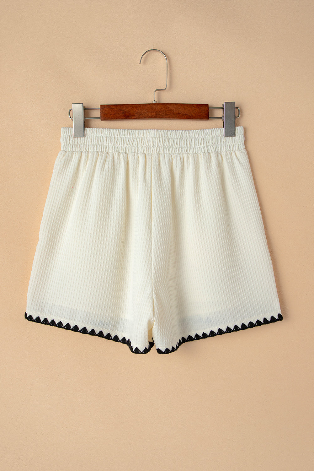 A lady wearing high-waisted white shorts with a textured waffle knit fabric and a contrasting ricrac detail along the hemline. They are dressed casually, with hands in their pockets, and are carrying a black and white handbag. The outfit is styled for everyday wear, suitable for pairing with shirts for a more dressy look.