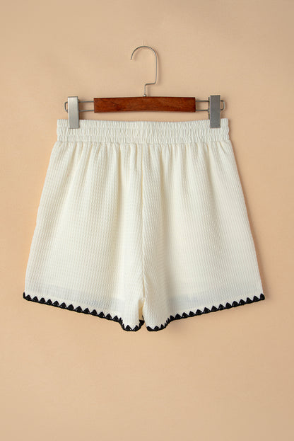 A lady wearing high-waisted white shorts with a textured waffle knit fabric and a contrasting ricrac detail along the hemline. They are dressed casually, with hands in their pockets, and are carrying a black and white handbag. The outfit is styled for everyday wear, suitable for pairing with shirts for a more dressy look.