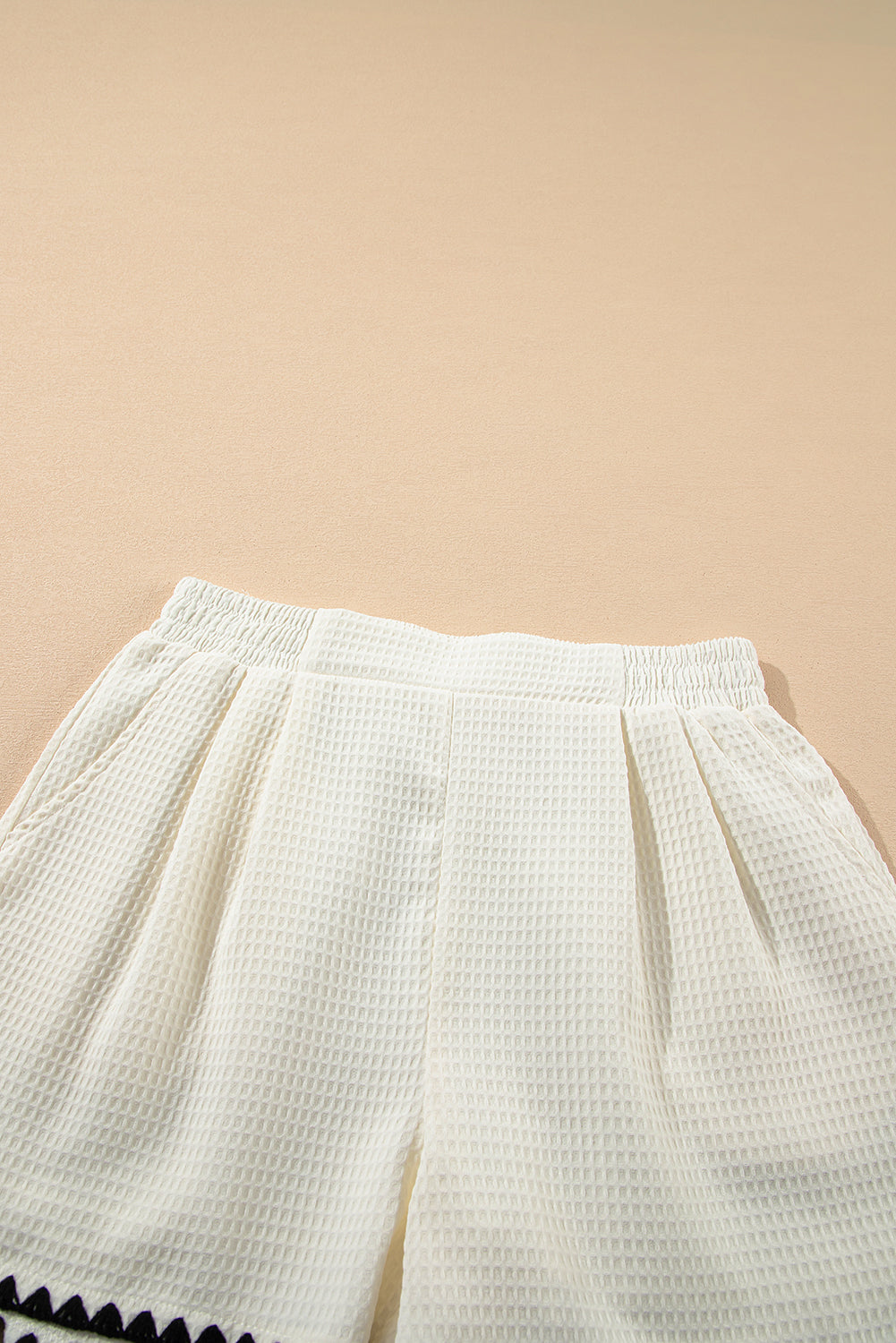 A high-waisted white shorts with a textured waffle knit fabric and a contrasting ricrac detail along the hemline. 