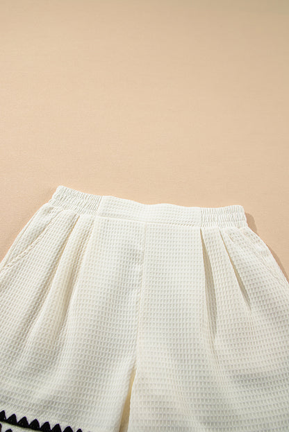A high-waisted white shorts with a textured waffle knit fabric and a contrasting ricrac detail along the hemline. 