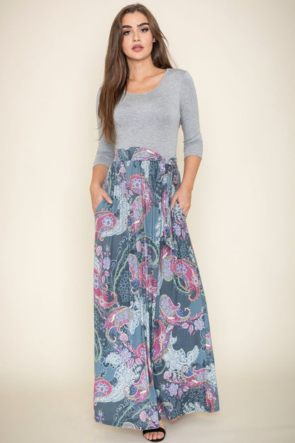 A woman with long brown hair is posing in a stylish outfit. She's wearing a white top with black stripes and a long white skirt with colorful circular patterns. She's got on tan heels and is touching her hair with one hand.