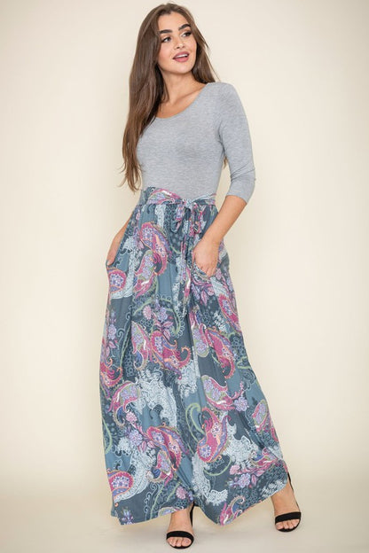 A woman with long brown hair is posing in a stylish outfit. She's wearing a white top with black stripes and a long white skirt with colorful circular patterns. She's got on tan heels and is touching her hair with one hand.