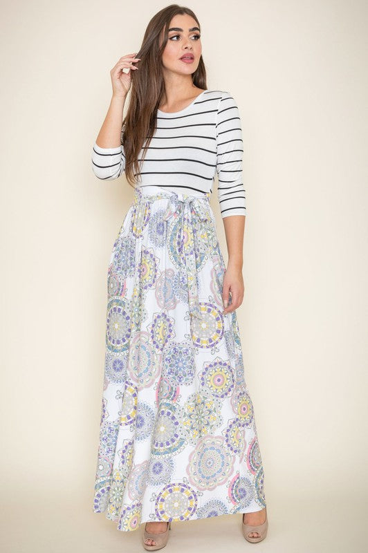 A woman with long brown hair is posing in a stylish outfit. She's wearing a white top with black stripes and a long white skirt with colorful circular patterns. She's got on tan heels and is touching her hair with one hand