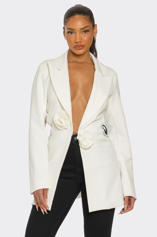  A lady posing in a sleek white blazer with floral details and a daring deep V-neckline, paired with black pants.