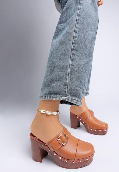Brown clogs with gold studs worn with light blue jeans on a gray background