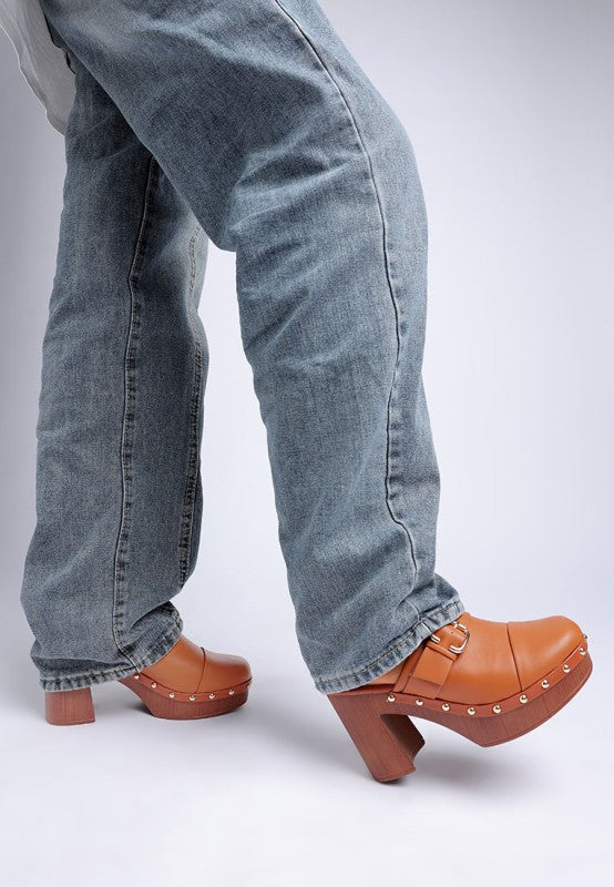 Person wearing blue jeans and brown clogs on a light gray background